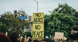 image from protest at head level of the crowd. A sign stands out from the rest, handwritten on a yellow board - No Justice No Piece